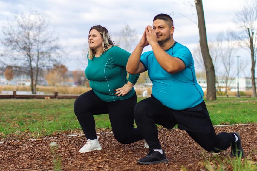 Two people wearing workout clothing exercising in the park. They are both kneeling down. The man has his hands brought together in front of him. The woman's hands are on her hips.
