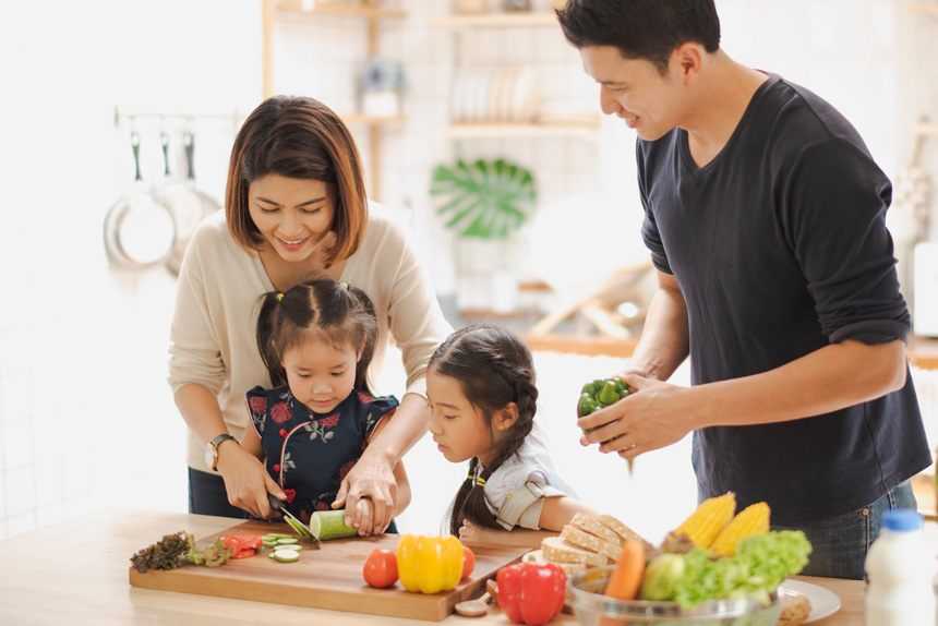 A family cooking together. A husband and wife supervise two young daughters while they cut vegetables.
